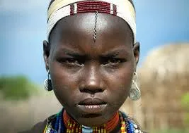 A young woman with a beaded headband, silver earrings and a beaded collar looks seriously at the camera.