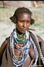 A young woman wearing many strands of white, blue, green and red beaded necklaces stands outdoors in a hide garment.