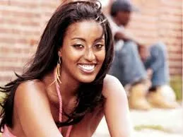 A smiling young woman with long dark hair and large earrings sits in front of a brick wall.