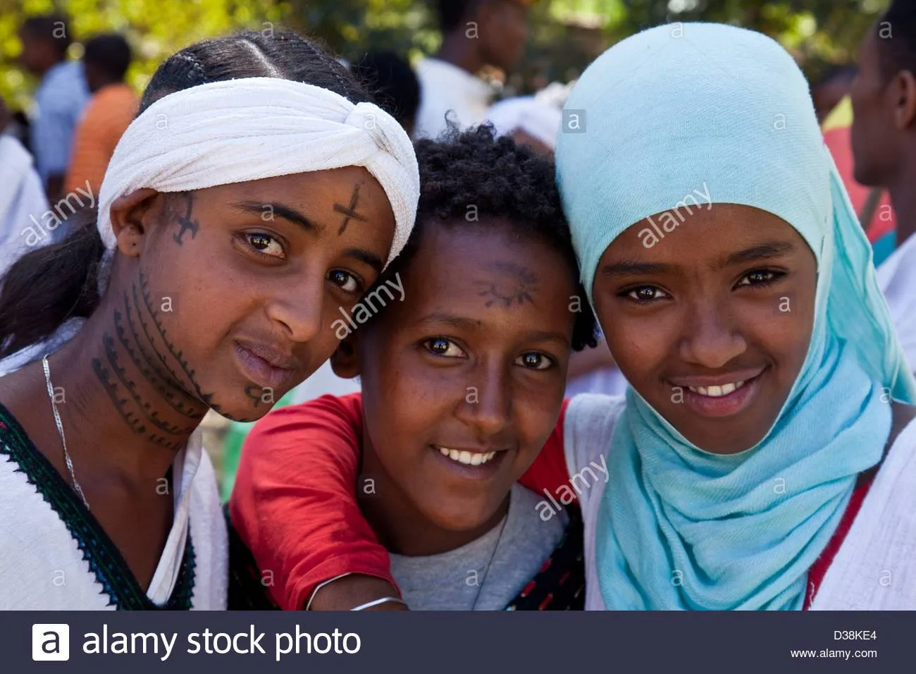Three smiling young people pose close together at an outdoor gathering; one wears a white head wrap and facial tattoos, another a light blue headscarf.