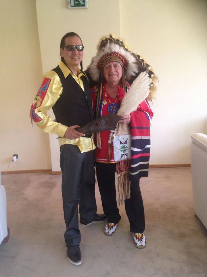 Koda Darcy in sunglasses, a gold shirt and black vest stands beside an elder in a feather headdress and red regalia holding a feather fan.