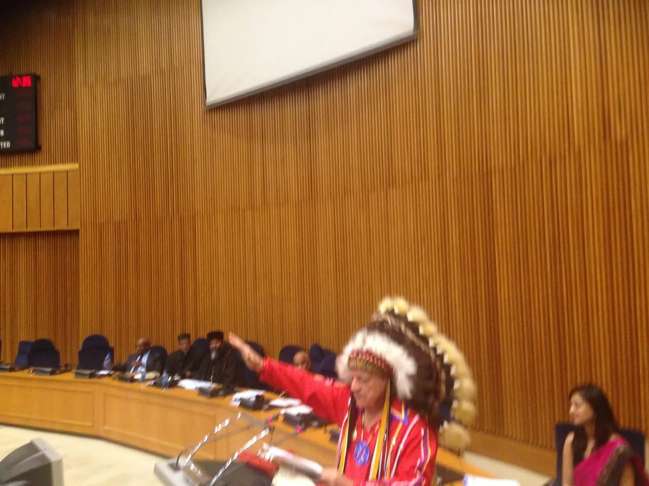 An elder in a feather headdress and red shirt raises his arm while speaking at a podium in a wood-paneled conference hall, with delegates seated behind.