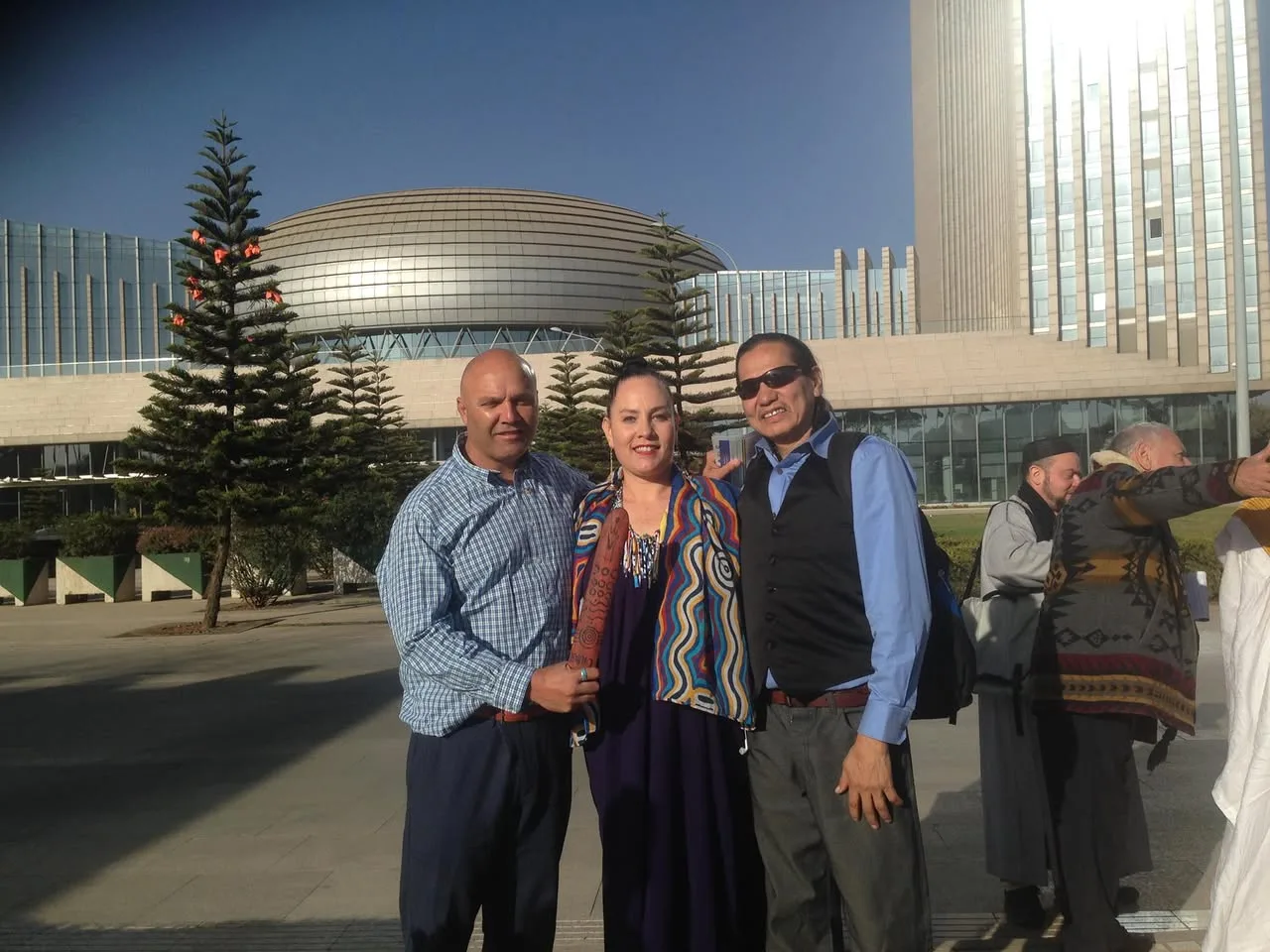 Two men and a woman in a colorful patterned shawl pose outdoors before the domed African Union Headquarters, others gathering nearby.