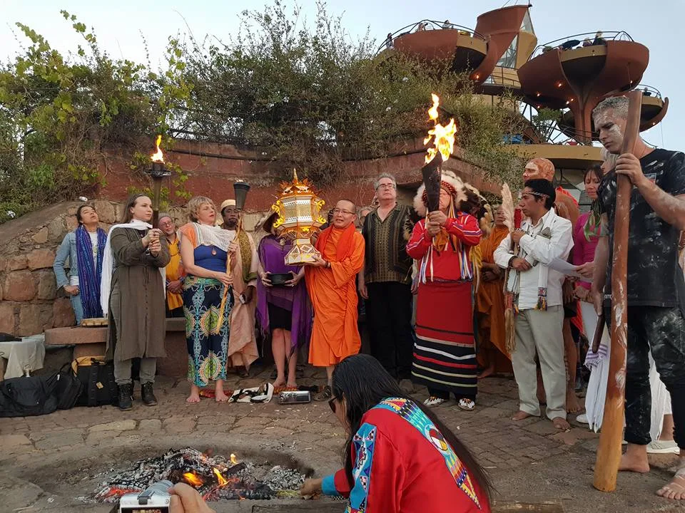 People of many faiths stand in a circle around a ceremonial fire at dusk, holding torches and a golden lantern, as a woman in a red ribbon shirt tends the fire.