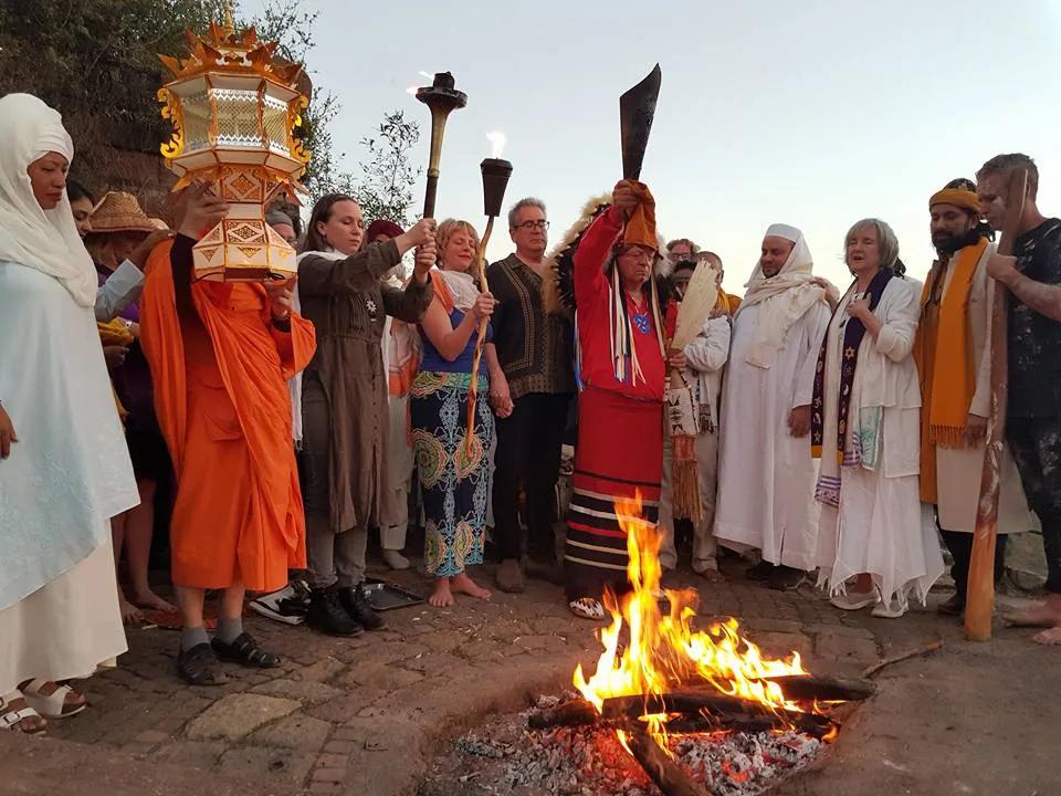 An elder in a feather headdress and red regalia raises a torch beside a fire, joined by a monk holding a lantern and people in white robes and shawls.