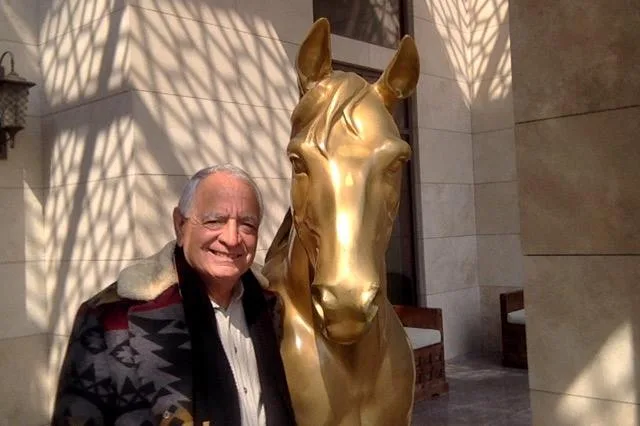 A smiling older man with white hair, wearing a patterned blanket coat with a sheepskin collar, stands beside a large golden horse statue in sunlight.