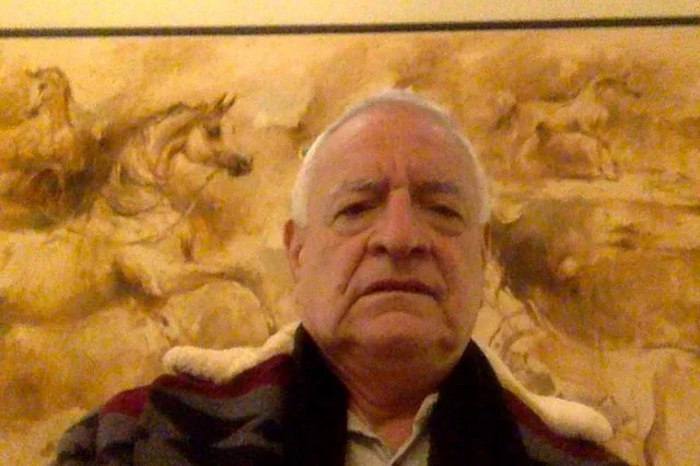 An older man in a patterned coat with a sheepskin collar sits in front of a golden mural of galloping horses.