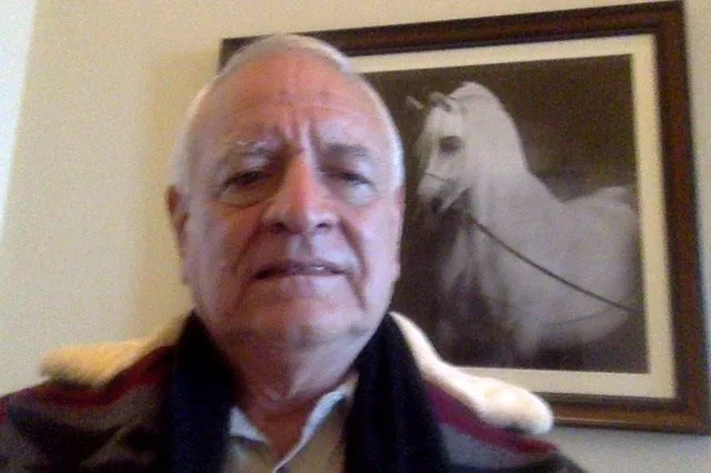 An older man in a patterned coat and dark scarf sits before a framed black-and-white photograph of a white horse.