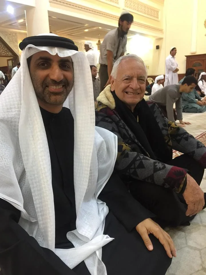 Phil Lane Jr. in a patterned blanket coat sits smiling on a mosque carpet beside a man in a white headdress and black robe, with worshippers behind them.