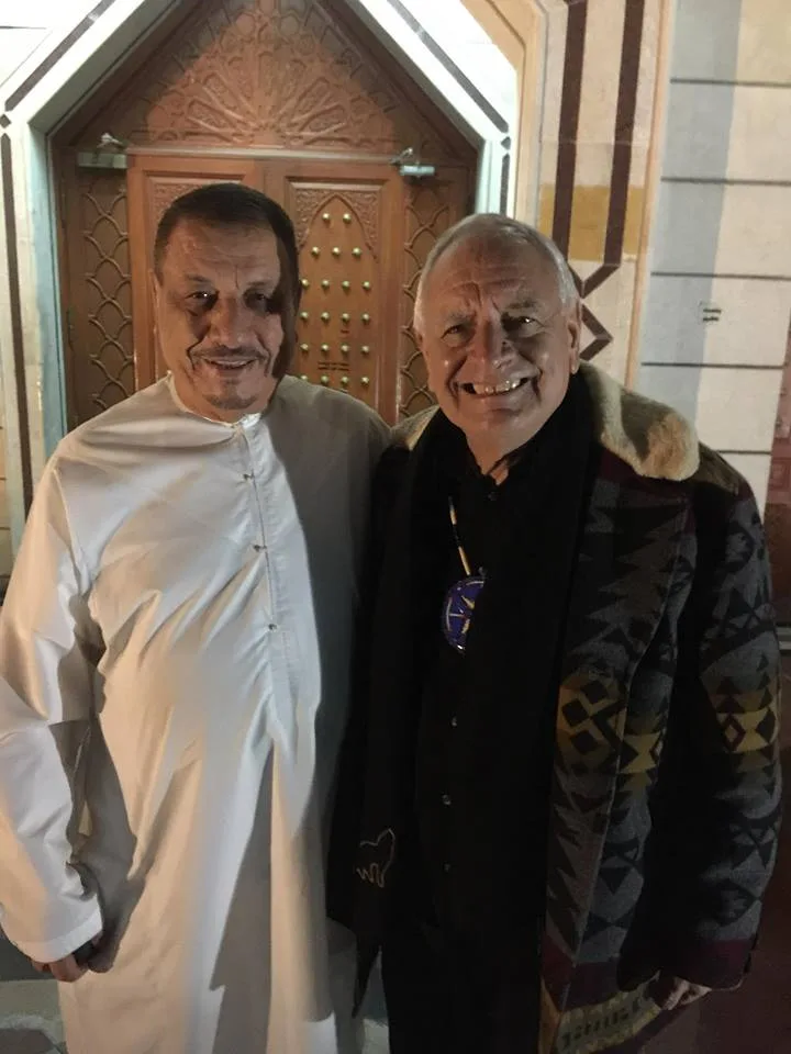 Phil Lane Jr. and a man in a white robe stand smiling with their arms around each other in front of an ornate carved wooden mosque door at night.