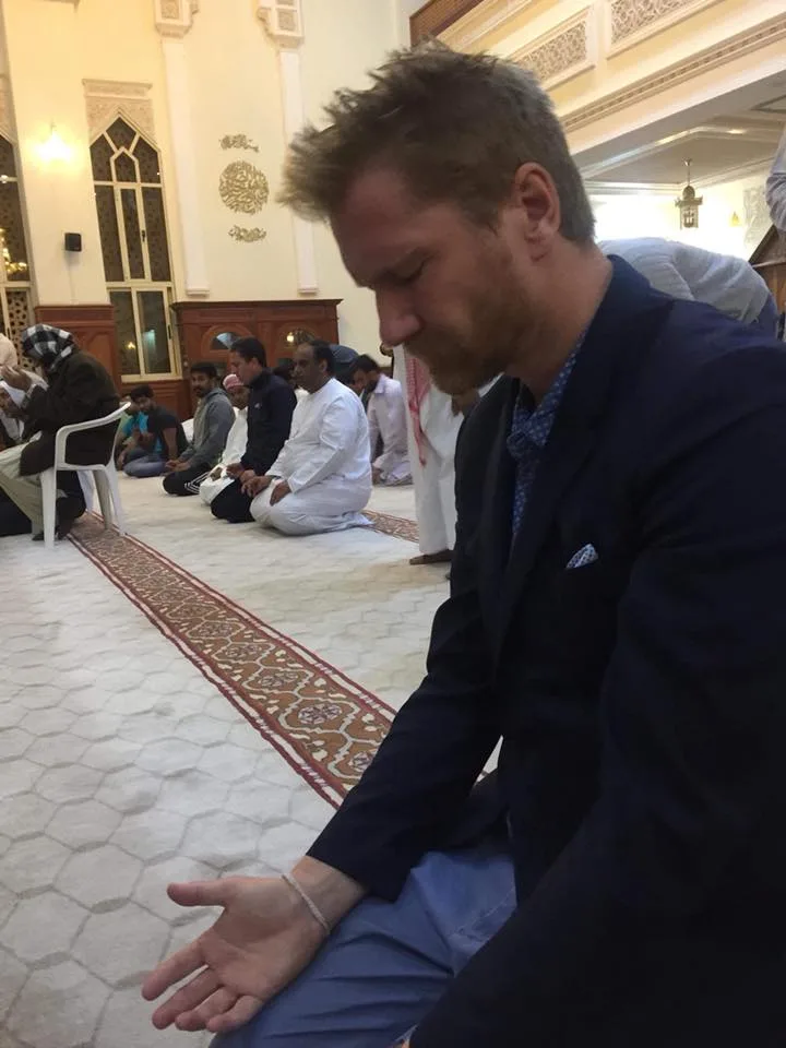 A bearded man in a navy blazer, believed to be Ben Bowler, kneels with eyes closed and palm open in prayer on a mosque carpet, with kneeling worshippers in a row behind him.
