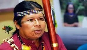 A Kichwa man from Sarayaku wearing a beaded headband, face paint markings and a seed necklace, holding a tall staff.