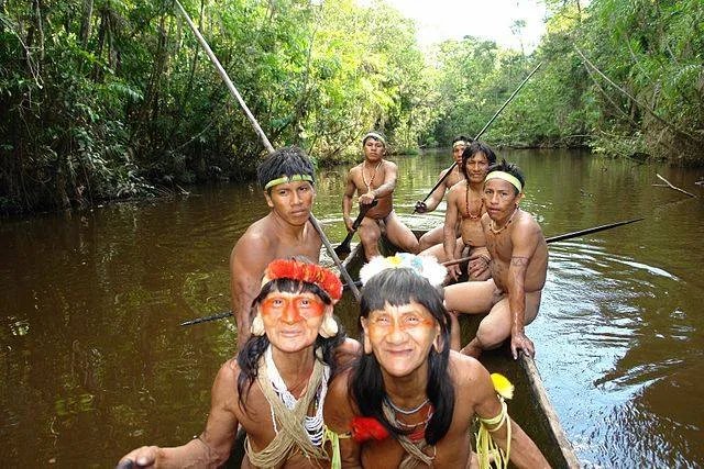 Indigenous women with orange face paint and feathered headpieces sit with men holding long paddles in a dugout canoe on a jungle river.