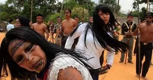 Indigenous women with long black hair and face paint lead a group of people walking on a dirt path, some holding staffs.
