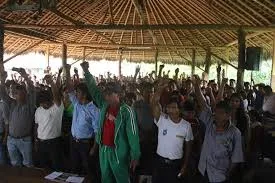 A large gathering of people under a thatched-roof meeting house, many raising their fists together.
