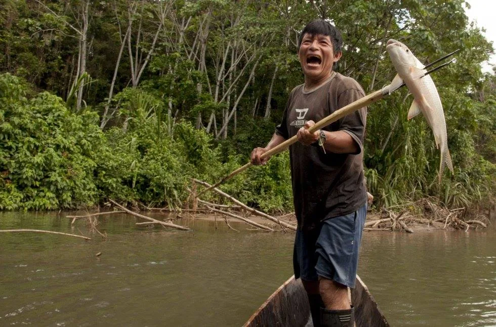 A smiling man standing in a dugout canoe on a river holds up a fish speared on a long wooden pole, with forest behind him.