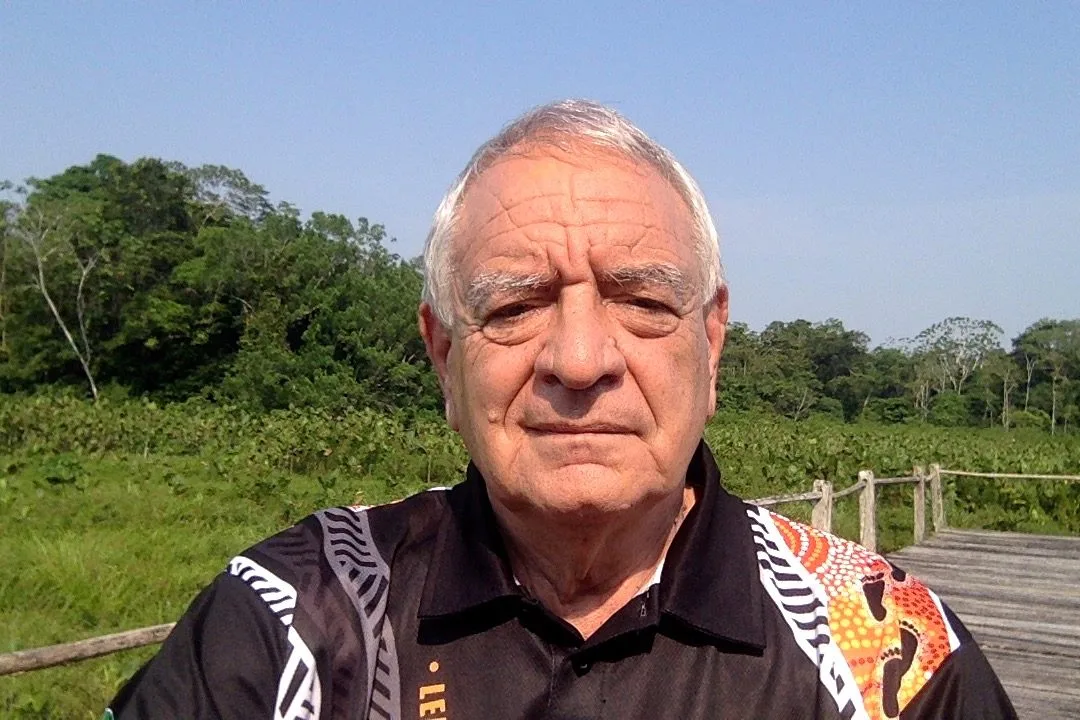 A white-haired man in a black shirt with Aboriginal-style dot art stands on a wooden boardwalk before green wetland and forest under a clear sky.