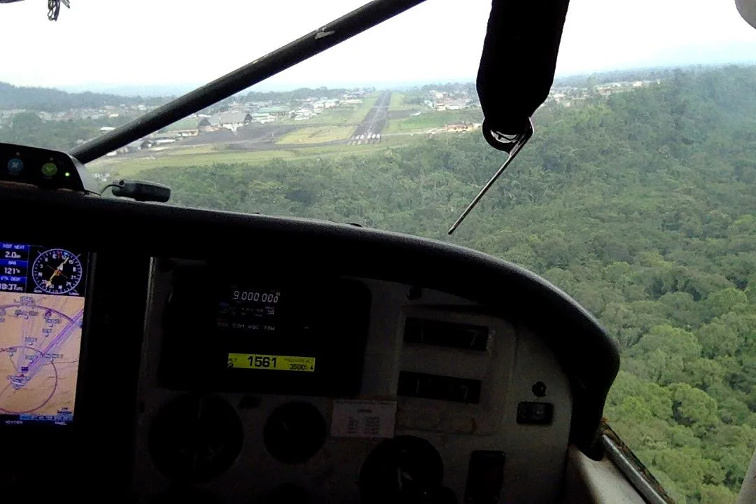 View from a small plane cockpit, with instruments and a GPS screen, approaching a runway and town beyond dense green forest.