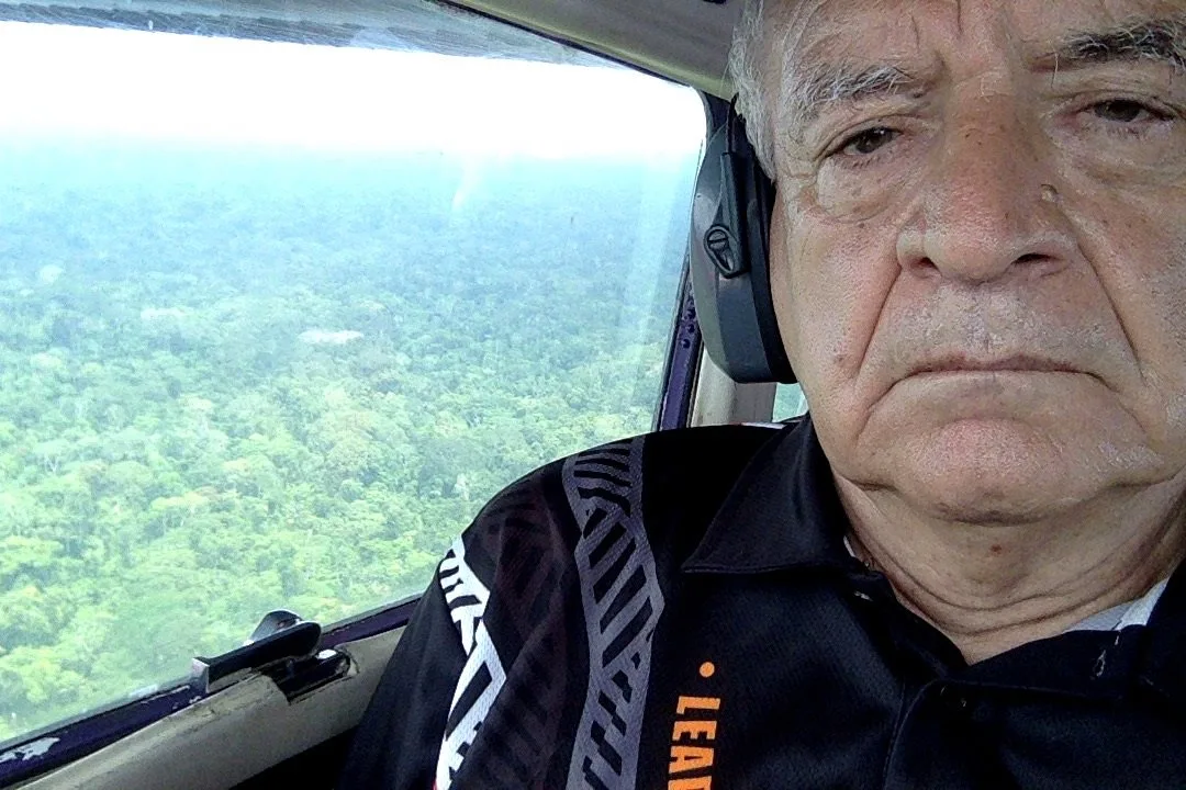 A white-haired man wearing a headset sits by a small plane window, with vast green Amazon rainforest stretching below.