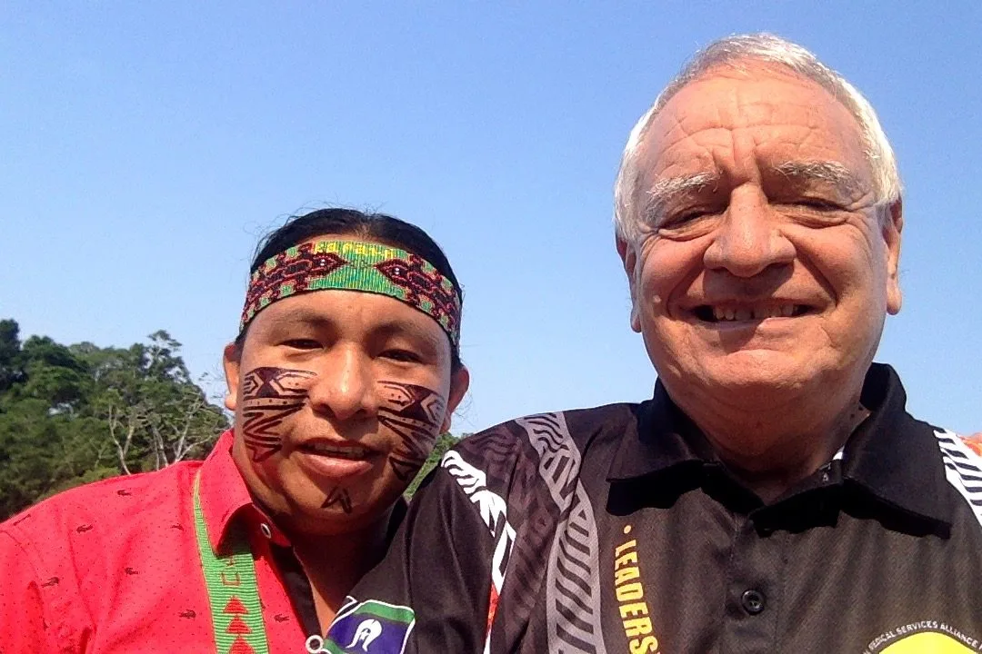 Selfie of Phil Lane Jr. and Cristobal Mukucham, who wears a beaded headband and red face paint designs, smiling under a blue sky with rainforest trees behind.