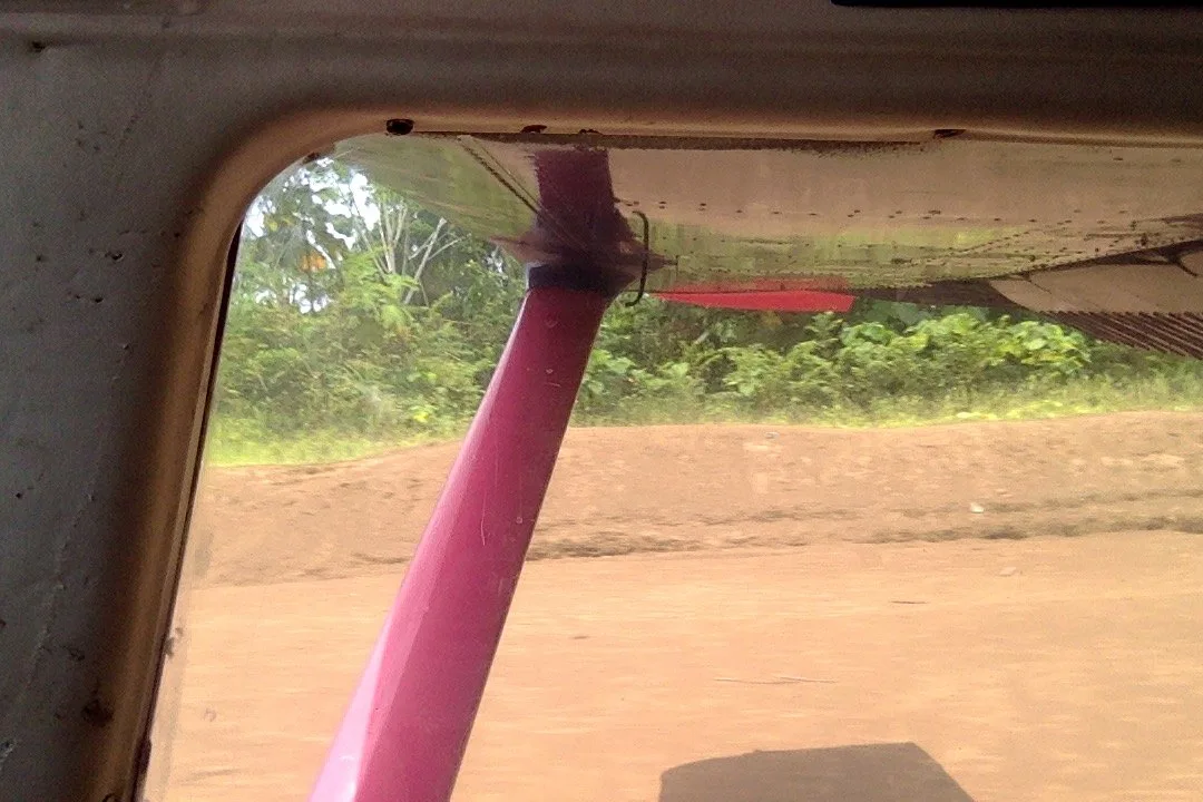 View from inside the small plane's window of a pink wing strut and a dirt runway lined with green jungle.