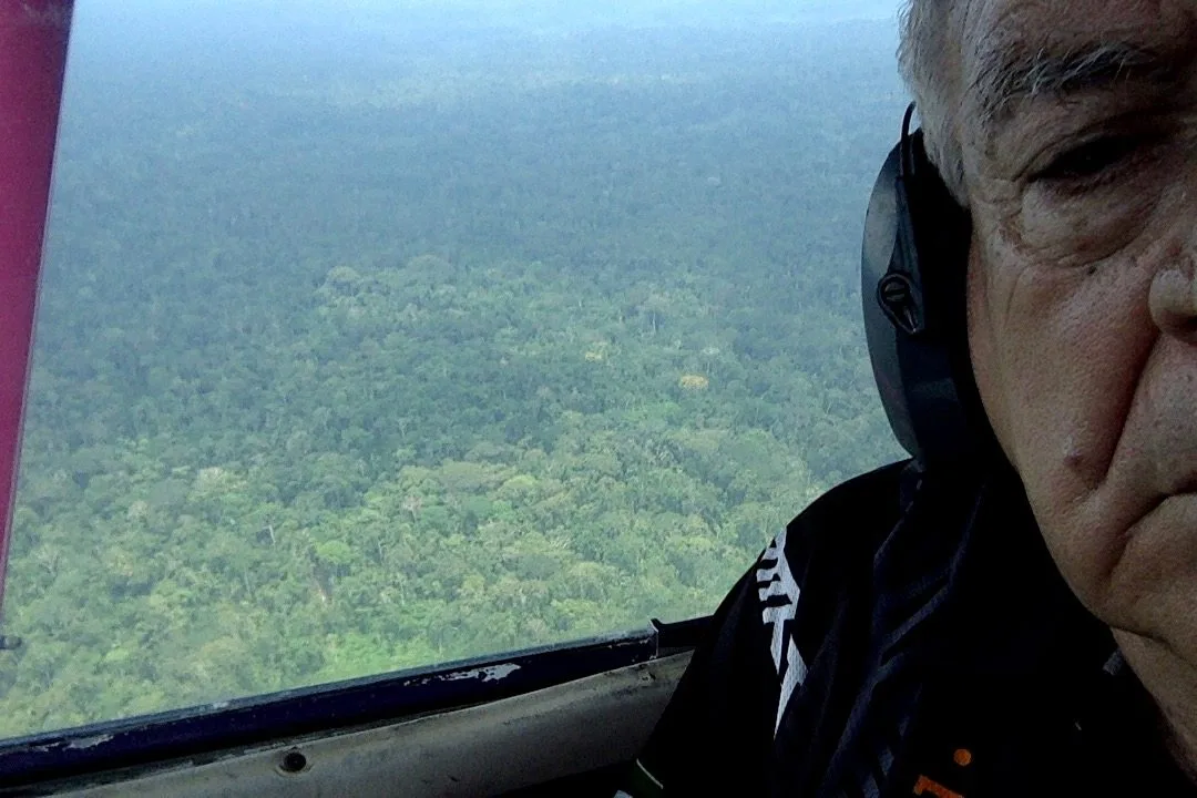 A grey-haired man wearing headphones takes a selfie in the plane as unbroken Amazon forest stretches below.