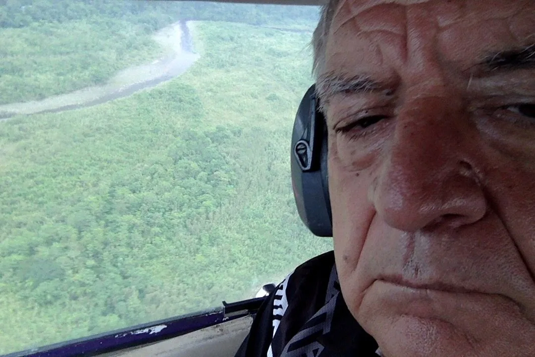 Close selfie of a grey-haired man in headphones in the cockpit, with green forest and a winding river visible below.