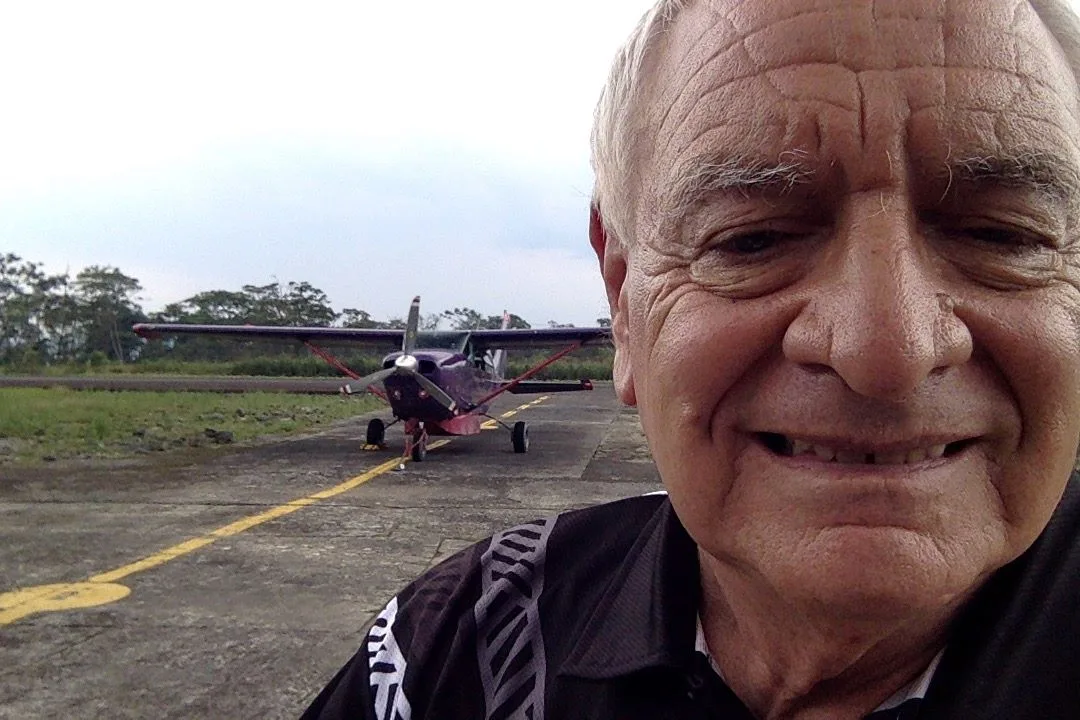 A smiling grey-haired man takes a selfie on a paved runway in front of the small purple plane.
