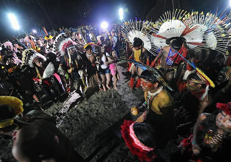 A nighttime gathering of Indigenous people in feathered headdresses, body paint and beads, dancing and singing together under bright lights.