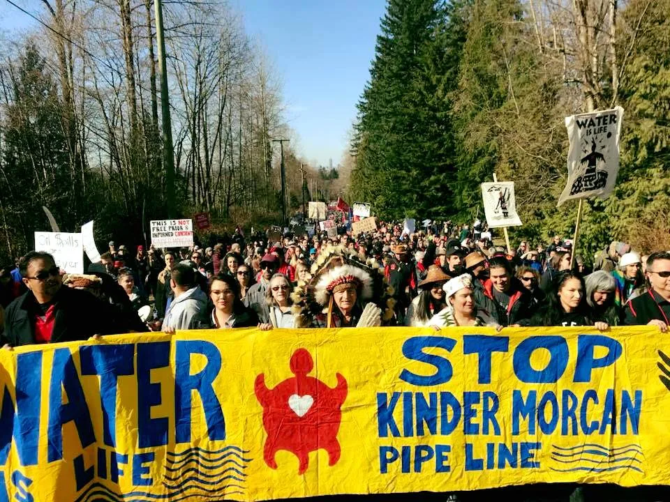 A large crowd marches down a tree-lined road behind a yellow banner reading "Water Is Life" and "Stop Kinder Morgan Pipeline," led by an elder in a feather headdress.