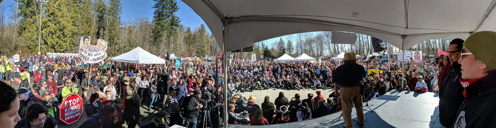 Panoramic view from a stage tent of a man in a hat addressing a vast crowd seated and standing in a forest clearing, holding signs against the Kinder Morgan pipeline.