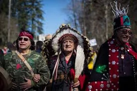 Three Indigenous elders march outdoors, the centre elder in a feathered headdress, the others in a camouflage jacket and a red and black regalia with a tall hat.