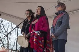Three Indigenous women stand on a stage under a tent; one in a red shawl sings into a microphone with a hand on her heart while another holds a hand drum.