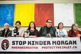 Indigenous leaders sit at a press conference table behind a banner reading "Stop Kinder Morgan, Kwekwecnewtxw, protecttheinlet.ca" before a colourful painted backdrop.
