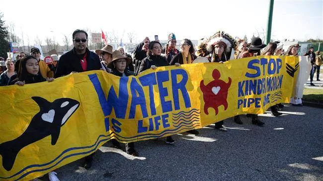 Marchers carry a long yellow banner reading "Water Is Life" and "Stop Kinder Morgan Pipeline," decorated with an orca and a red turtle, joined by an elder in a feather headdress.