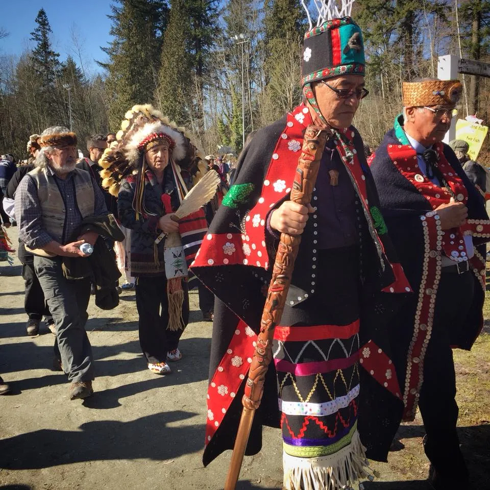 Elders in ceremonial regalia walk along a forest path, one holding a carved staff and wearing a red and black button blanket, followed by an elder in a feathered headdress.
