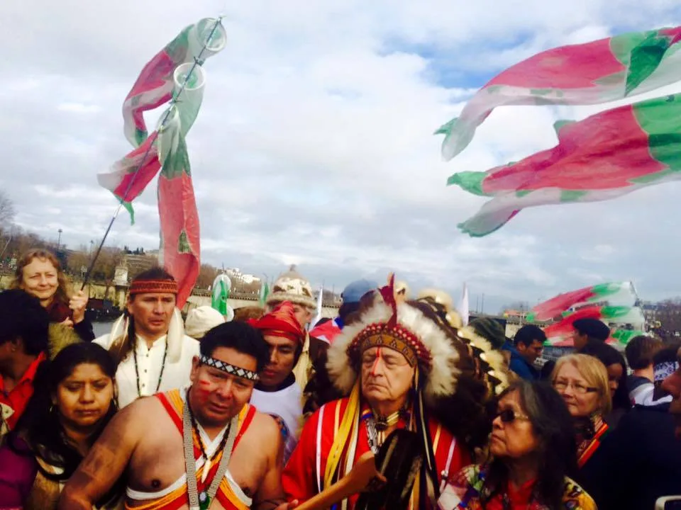 A crowd of Indigenous people in traditional regalia, including an elder in a feathered headdress and face paint, gathers outdoors beneath red and green streamers.