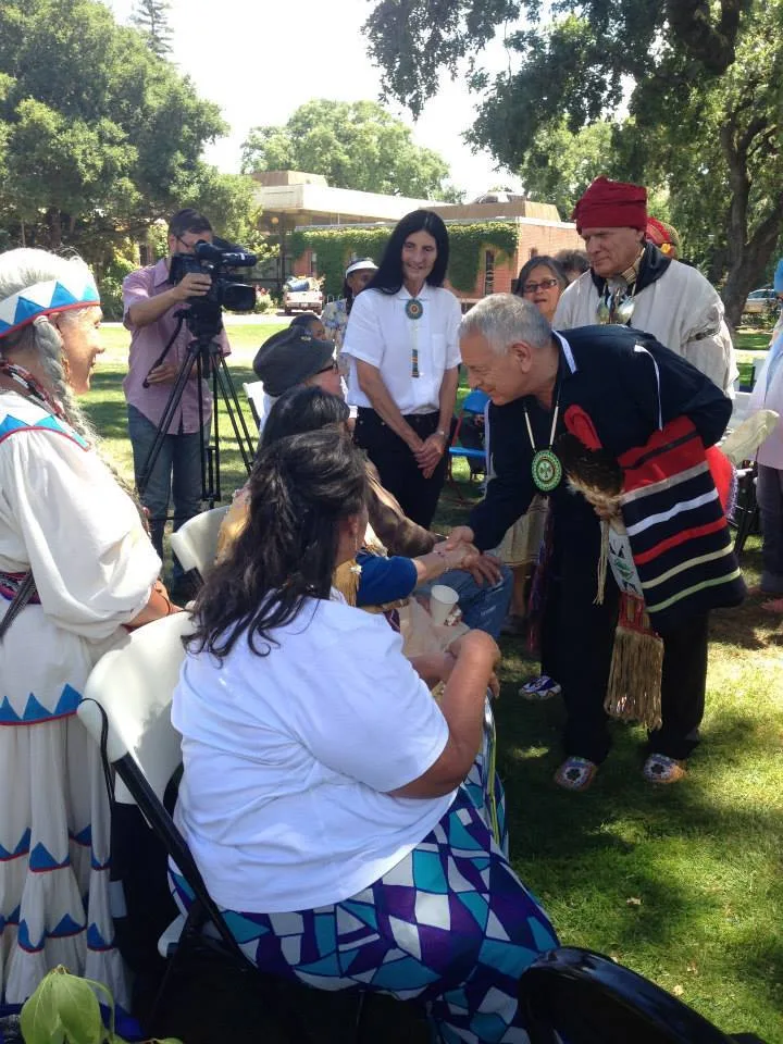 An elder in a dark ribbon shirt and beaded medallion leans to shake hands with seated women at an outdoor gathering on a lawn, as a camera operator films nearby.