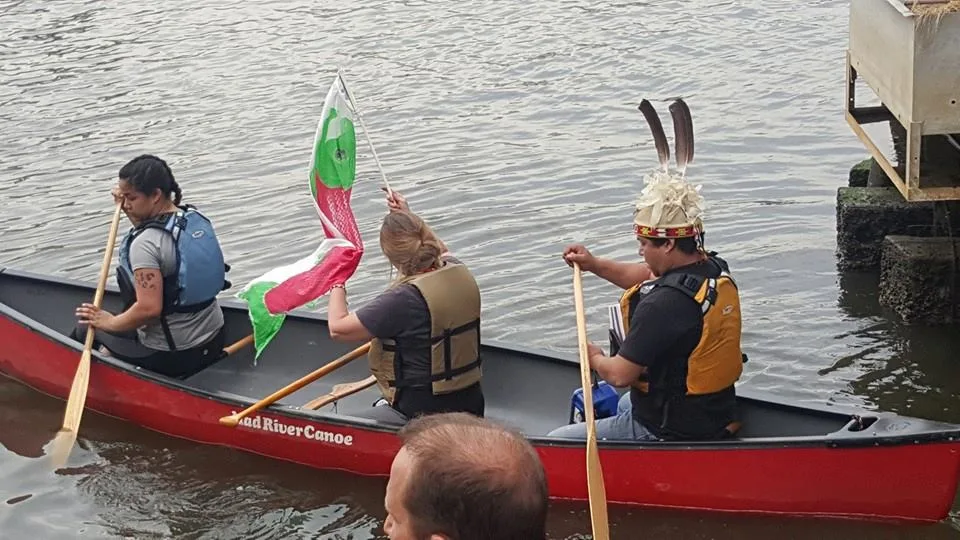 Three paddlers in life jackets in a red canoe on a river, one waving a green, white and red flag and one wearing a feathered headdress.