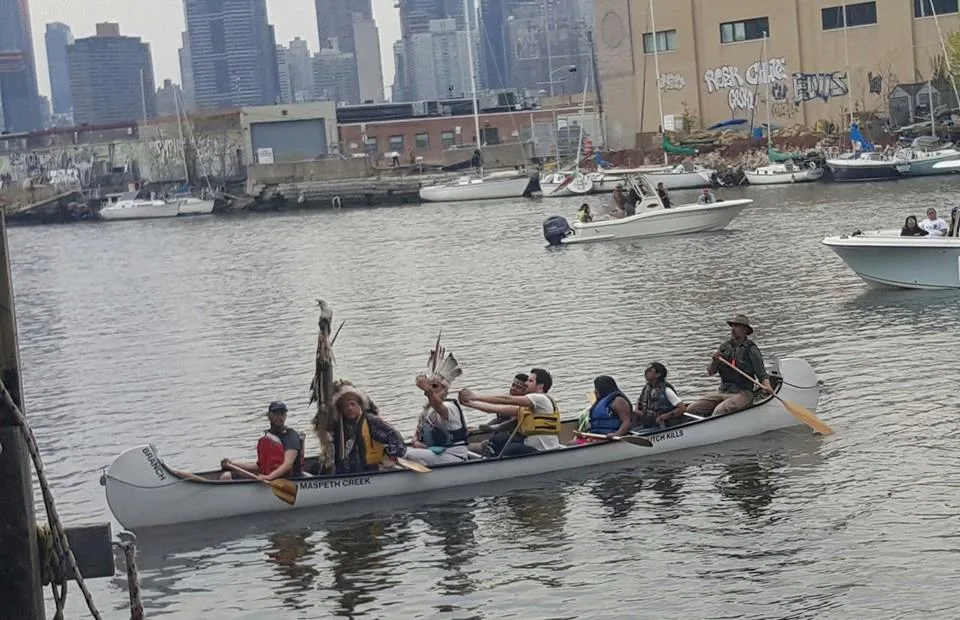 A long white canoe carrying paddlers, some in feathered headdresses and one holding a ceremonial staff, on the water with boats and city skyscrapers behind.