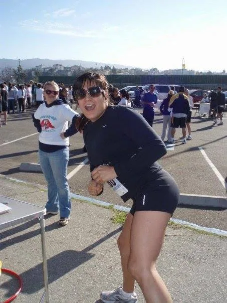 A smiling young woman in sunglasses and black athletic wear with a race number pinned on, posing playfully in a parking lot crowded with runners and spectators.