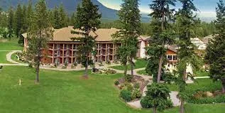 A multi-storey lodge with balconies set among tall evergreens and green lawns, with a forested mountain behind.