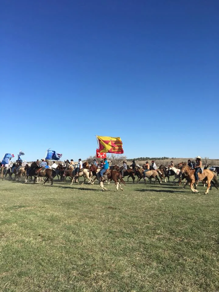 Many riders on horseback cross a grassy plain under a clear blue sky, carrying flags including a large red and yellow flag.