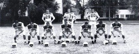 Black-and-white team photo of a high school football offense on a grass field: seven linemen crouched in stance with helmets on the ground before them, four backs standing behind.
