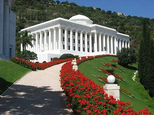 A white neoclassical building with columns and a dome on a green hillside, approached by a gravel path lined with red flowers.
