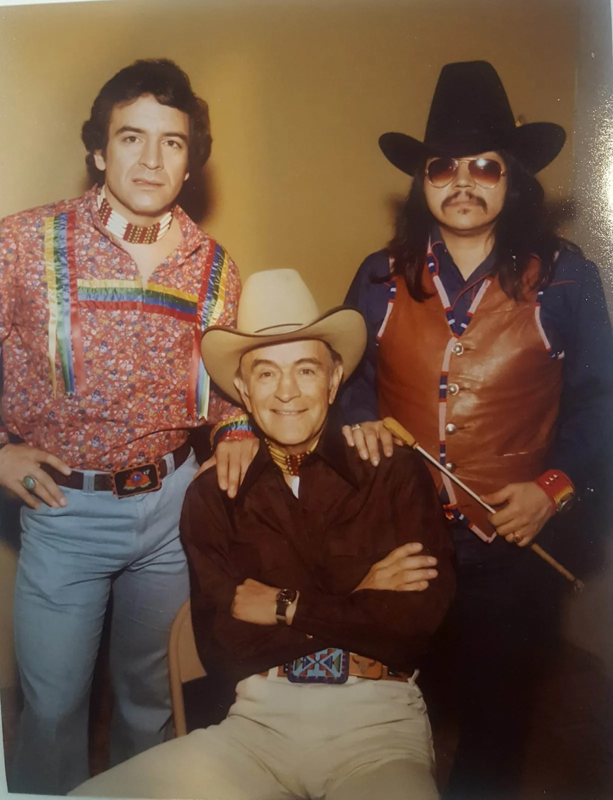 Phil Lane Jr. in a ribbon shirt and bone choker stands beside his smiling father, seated in a cowboy hat, and Koda George in a black hat, sunglasses and beaded leather vest.