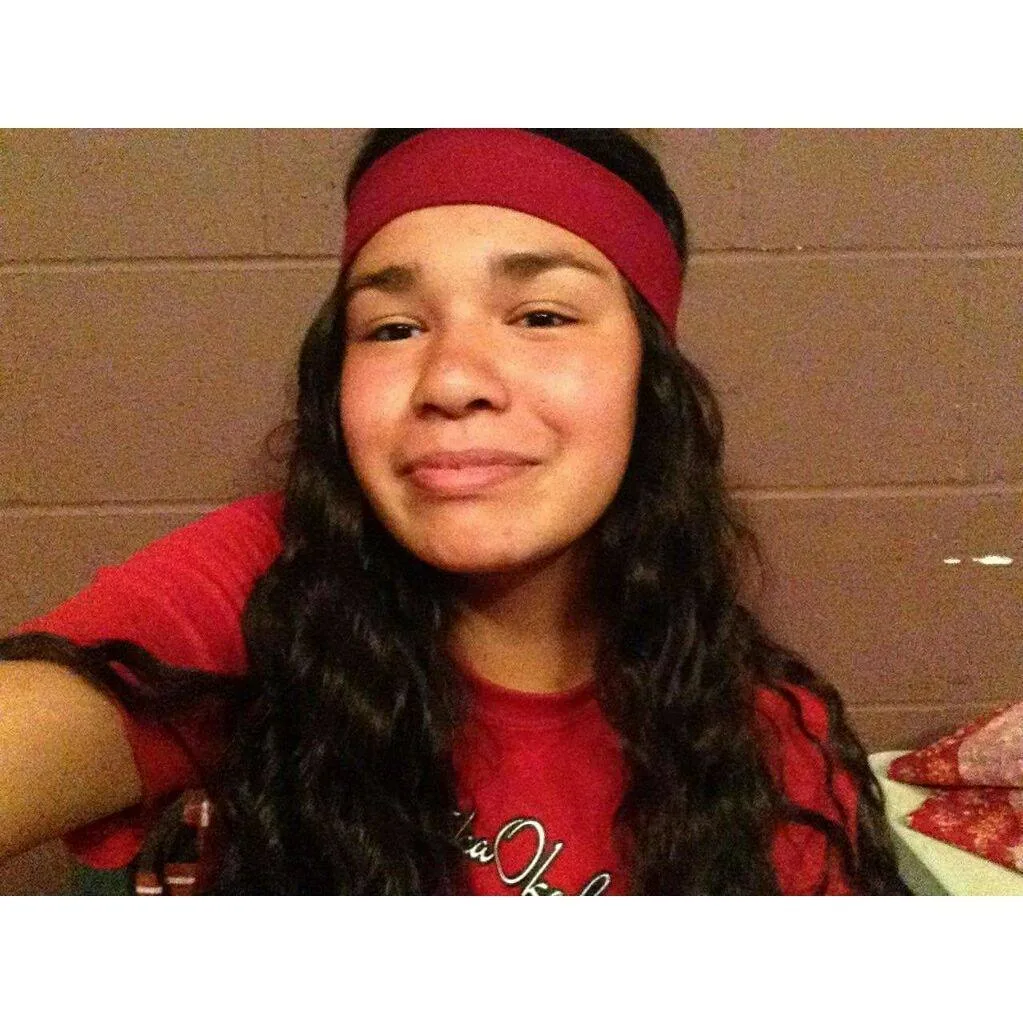 Smiling young woman with long dark wavy hair and a red headband, wearing a red t-shirt, taking a selfie against a brown wall.
