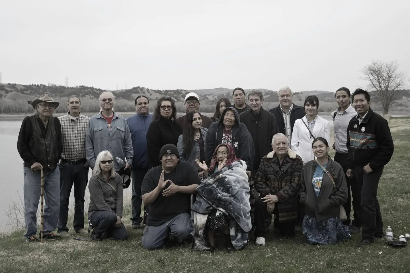 Large group of Indigenous elders, youth and supporters posing outdoors beside a lake with rolling hills behind, an elder wrapped in a blanket seated at center.