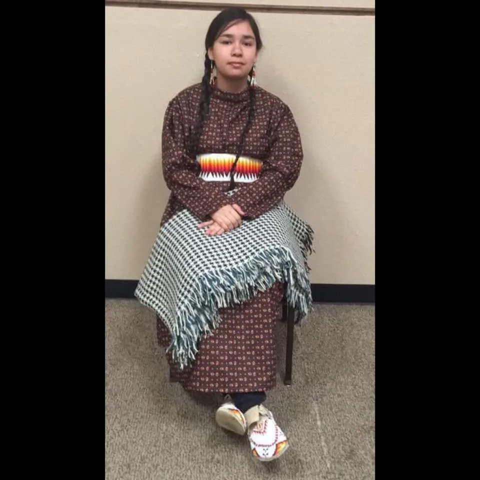 Young woman with long braids seated on a chair in a brown print dress with a beaded belt, a fringed shawl across her lap, and beaded moccasins.