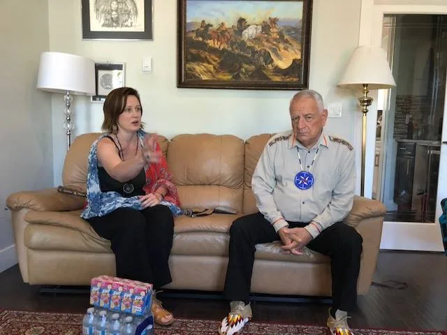 A woman in a colorful scarf gestures as she speaks, seated on a tan leather sofa beside Phil Lane Jr., an elder in a light shirt and beaded moccasins, in a living room with Western paintings.